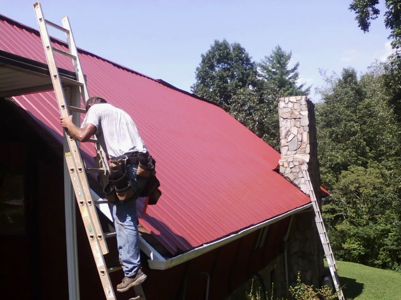 Roofer on a ladder beside a red metal roof and stone chimney — Soffit & Fascia Repair by Roofing Brothers in North Bloomfield